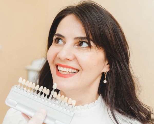 Smiling woman comparing her teeth to a veneer shade guide